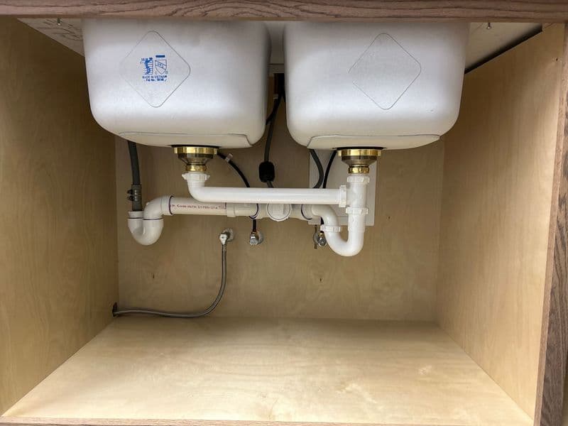 Under-sink plumbing with double sinks and PVC pipes in a wooden cabinet interior.