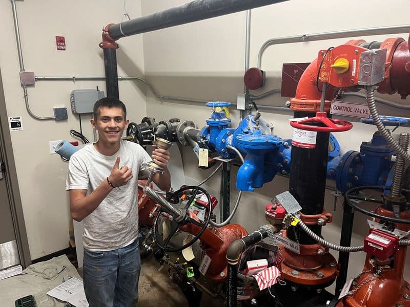 Young technician thumbs up beside industrial water control valves and pipes in a workshop.
