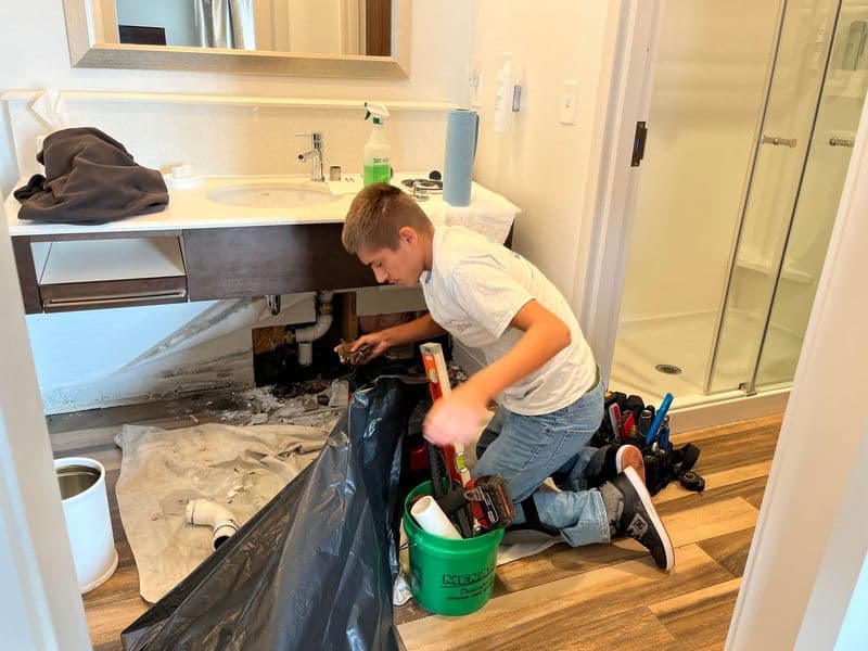 Boy fixing a sink under a bathroom cabinet, using tools and a plastic sheet for protection.