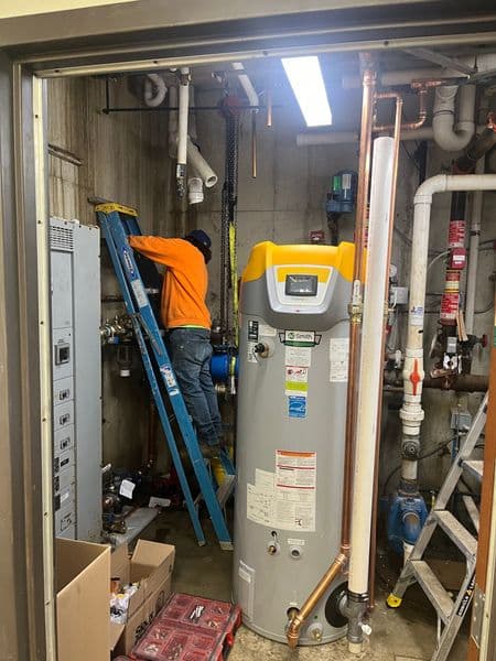 Technician repairing plumbing system, using ladder near water heater in mechanical room.