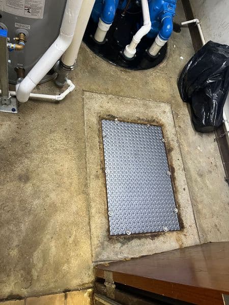 Metal access cover on concrete floor, surrounded by plumbing and a black trash bag.