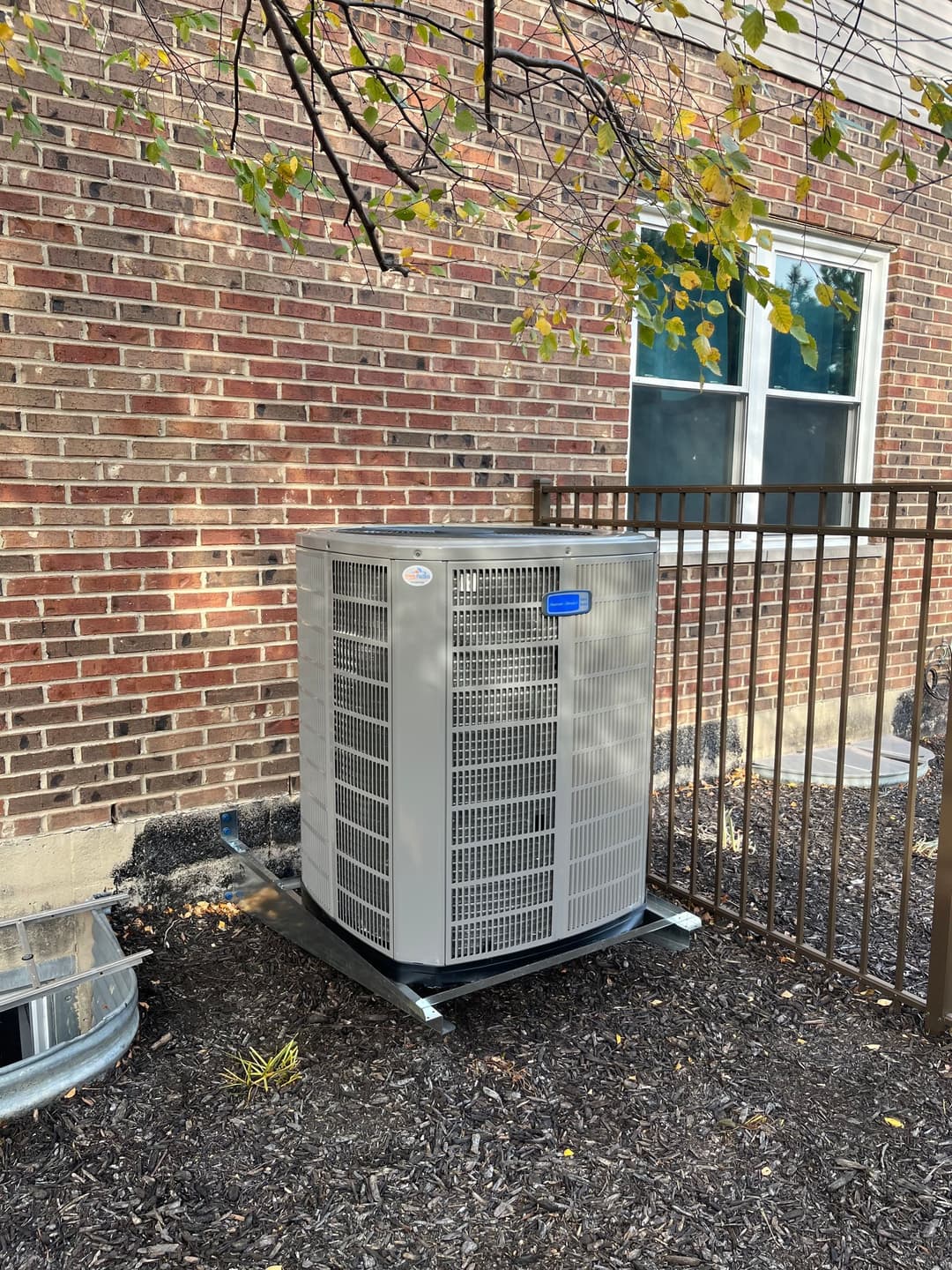 Air conditioning unit installed near a brick building with a fenced area and vegetation.