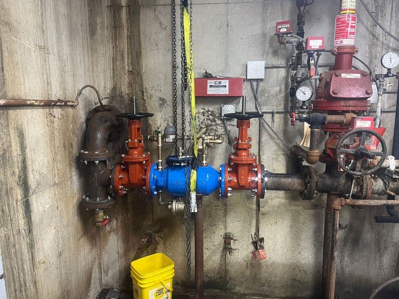 Industrial water valve assembly with pressure gauges and safety equipment in a utility room.