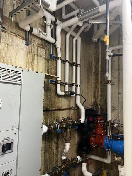 Complex plumbing installation with white pipes, valves, and electrical panel against stained wall.