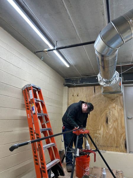 Worker using plumbing tools near a ladder and ventilation duct in a construction area.