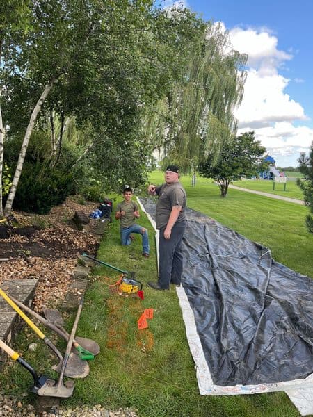 Two landscapers working on garden renovation with tools and a tarp in a grassy area.