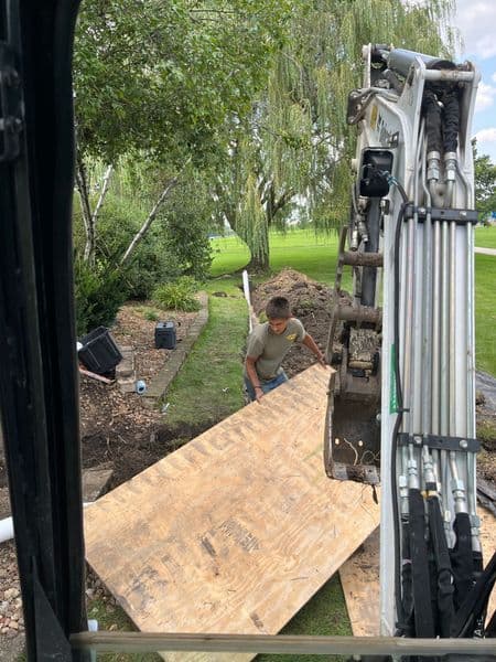 Construction worker laying plywood on a landscape site, with excavation visible in the background.