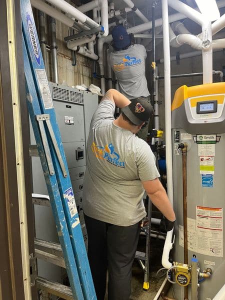 Technicians installing plumbing equipment in a mechanical room with a ladder.